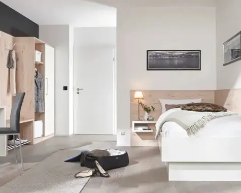Helles Schlafzimmer mit weissem Bett vor Holzrückwand und Nische mit Lampe, links offener Garderobenschrank und Stuhl, vorn geöffneter Koffer auf Teppichläufer.